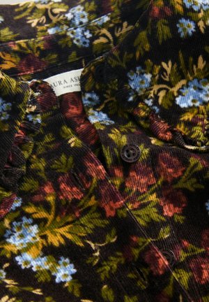 Knapperet corduroy-stof med multikolor floral tryk i grønne, blå, røde og gule toner, med synlige knapper og mærketekst.