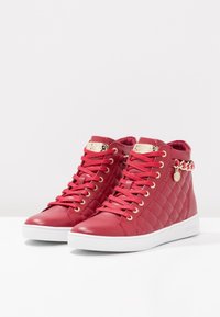 Red high-top sneakers made of quilted faux leather, featuring gold eyelets, laces, a gold logo plate, and a decorative chain accent. White sole.