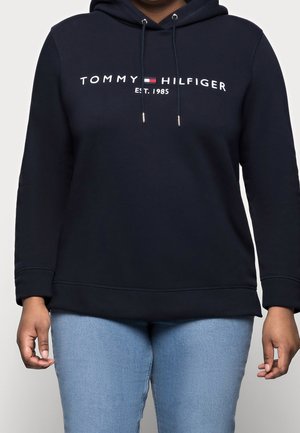 Osoba ubrana w granatową bluzę z kapturem Tommy Hilfiger z logo i napisem „Est. 1985”, w połączeniu z jasnoniebieskimi jeansami.