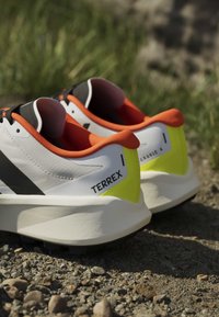 Chaussures de trail blanches et jaunes avec une doublure orange portant l'étiquette "TERREX GRAVIC" reposant sur un sol rocheux en plein air.