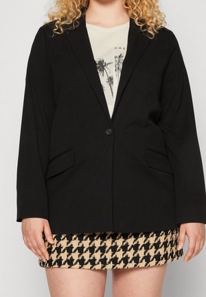 Personne portant un blazer noir sur une chemise blanche avec un imprimé de palmiers, associé à une jupe à motif pied-de-poule noir et beige.