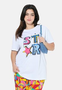 Camiseta blanca de algodón con texto gráfico en negrita y colorido, y diseños de estrellas. Combinada con pantalones de patrón multicolor y un bolso azul.