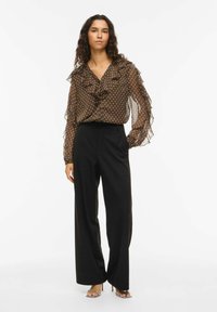 Blusa marrón con lunares y detalles de volantes, combinada con pantalones anchos negros y sandalias plateadas de tacón. Ella tiene el pelo largo y rizado.