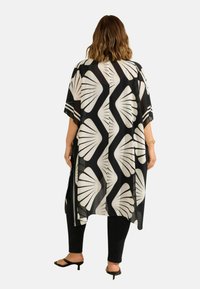 Läpinäkyvä musta kimono, jossa on näyttävät valkoiset lehtikuviot, lyhyet hihat ja avoimet sivut, kerroksittain mustien housujen ja korkokenkien päällä.