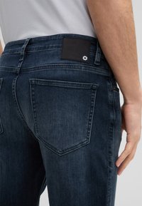 Dunkelblaue Denim-Jeans im Slim-Fit-Stil mit glatter Textur und zwei Gesäßtaschen. Ein schwarzes Leder-Patch mit Logo ist am Bund sichtbar.
