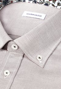 Gros plan sur une chemise blanche à petits points foncés, col boutonné, avec une étiquette visible de la marque Seidensticker à l'intérieur de l'encolure.