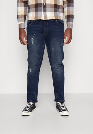 Jeans in denim blu scuro con dettagli strappati, taglio dritto, cinque tasche, indossati con sneakers alte nere e una camicia a quadri.