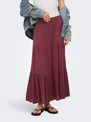 Naine kannab pikka, kihilist burgundia seelikut, valget topi, denimjope õlgadel ning musti flip-flop-susse valgel taustal.