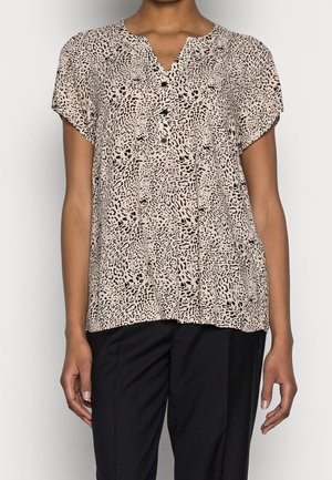 Femme portant un chemisier beige à manches courtes avec des taches abstraites noires et des boutons à l'avant, associé à un pantalon noir, debout devant un fond uni.