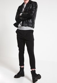 Black leather biker jacket over a gray sweatshirt, paired with black pinstripe trousers and black lace-up boots. Model stands with arms crossed.