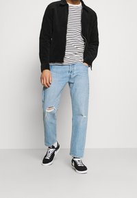 Svart kordflöjlsjacka, randig vit och marinblå T-shirt, ljusblå slitna jeans och svarta sneakers med vita accenter.