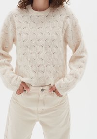 Krämfärgad stickad tröja med ett strukturerat vågmönster; avslappnad passform och ribbade ärmslut. Matchad med ljusbeiga högmidjade jeans med knappstängning.