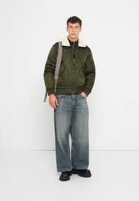 Veste bomber vert olive avec un col en fausse fourrure crème, associée à un jean large bleu clair et des chaussures noires à semelles épaisses. Fermetures éclair argentées visibles.