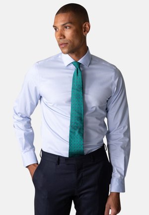 Mann iført lyseblå skjorte, blågrønt mønstrete slips og mørke bukser, ser til siden med én hånd i lommen.