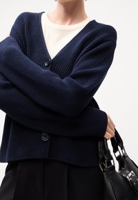 Námořnický pletený cardigan s výstřihem do V, který má žebrovanou strukturu a dva knoflíky, nošený přes krémový top, s černou kabelkou drženou v ruce.