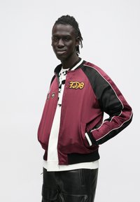 Burgundia satiinist bomberjakk mustade detailide, valge paelte ja tikitud embleemidega; kantud mustrilise särgi ja mustade pükste peal.