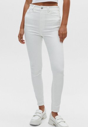 Femme portant un jean skinny blanc taille haute et des mocassins blancs épais, debout devant un fond blanc uni.