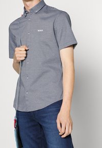 Szara koszula na krótki rękaw z guzikami, o fakturowanym wzorze i małym logo „BOSS” na piersi. Noszona z niebieskimi jeansami.