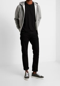 Veste à capuche grise zippée portée sur un t-shirt noir, associée à un pantalon noir retroussé et des baskets grises avec des lacets blancs. Design minimaliste et look décontracté.