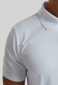 Valkoinen polo-paita, jossa on klassinen kaulus ja lyhyet hihat, valmistettu pehmeästä kankaasta, jolla on sileä pinta ja minimaalinen ompeluysitys.