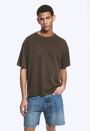 Jeune homme aux cheveux courts et bouclés, portant un t-shirt ample marron foncé et un short en jean bleu clair, debout avec une main dans la poche.