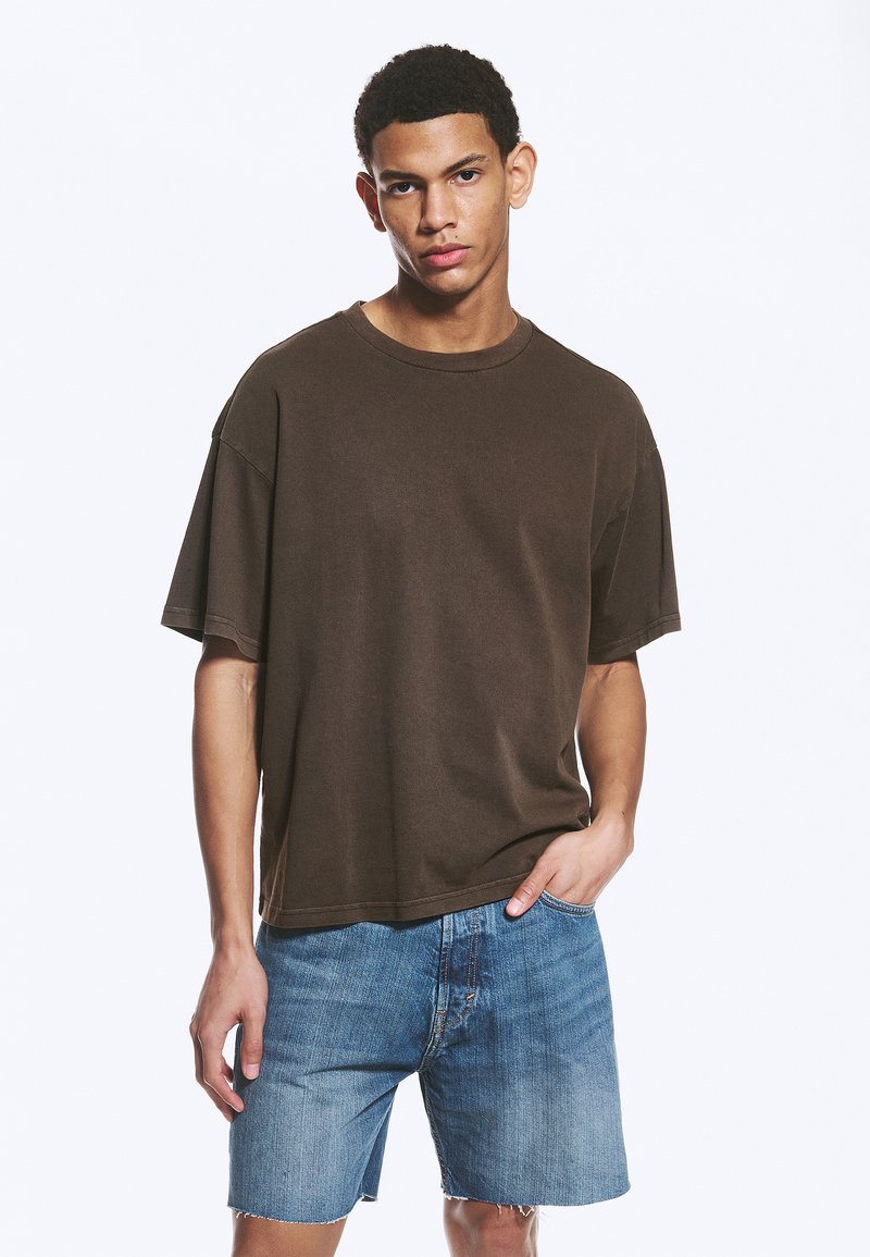 Jeune homme aux cheveux courts et bouclés, portant un t-shirt ample marron foncé et un short en jean bleu clair, debout avec une main dans la poche.