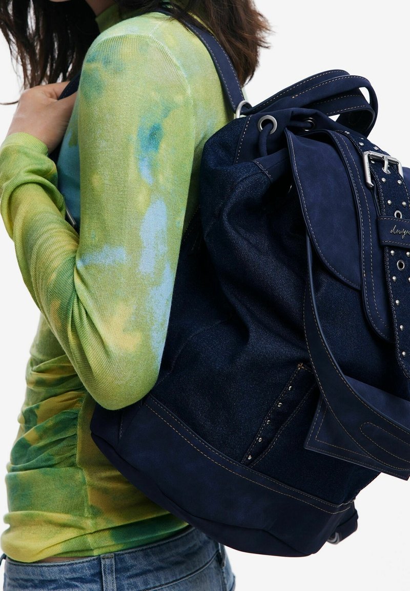Femme portant un t-shirt à manches longues tie-dye vert et bleu, avec un grand sac à dos en denim bleu marine et cuir porté sur une épaule.