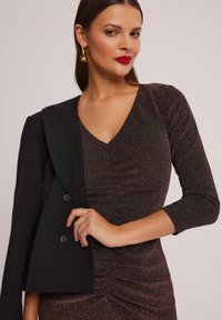 Długie rękawy, sukienka z głębokim dekoltem w kształcie V w ciemnej, błyszczącej tkaninie z marszczeniami, połączona z dopasowanym czarnym żakietem z okrągłymi guzikami.