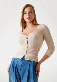 Beige ribbed cardigan with a V-neckline, featuring decorative buttons and a fitted design. Worn over high-waisted blue denim trousers.