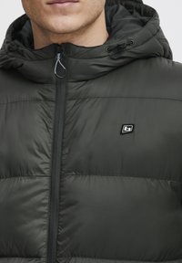 Mørkegrøn polstret jakke med lynlås og hætte. Har et lille logo på brystet og en glat, blank tekstur.