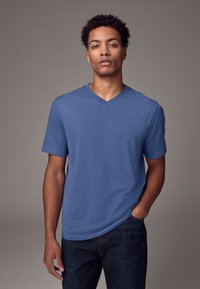 Modra v-neck majica iz mehkega, lahkega materiala; kratki rokavi in sproščen kroj, v kombinaciji s temno jeans hlačami, stoji sredi nevtralne ozadja.