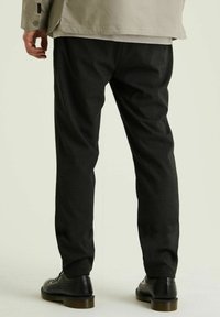 Czarne spodnie na miarę o fakturowanym splocie, prostym kroju i lekko zwężanych nogawkach, noszone z czarnymi sznurowanymi butami.