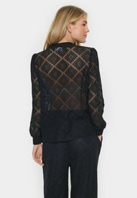 Top in pizzo nero con motivo a diamante, maniche lunghe e orlo a tinta unita in contrasto. La parte posteriore presenta dettagli trasparenti con una finitura testurizzata.
