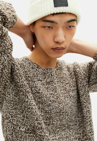 Maglione lavorato a maglia marrone e bianco con collo a giro, caratterizzato da un motivo testurizzato. Un berretto bianco con etichetta completa il look.