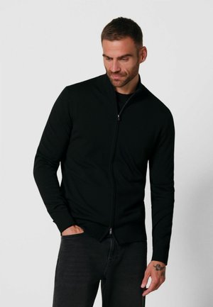 Uomo che indossa una giacca nera con zip e pantaloni scuri con una mano in tasca, guarda verso il basso sul lato sinistro su uno sfondo semplice.