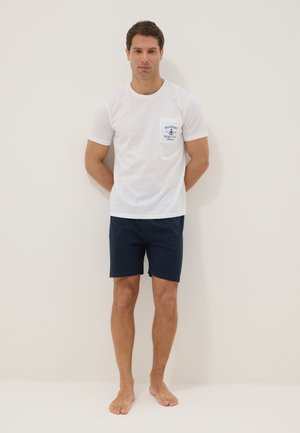 Homme debout pieds nus portant un t-shirt blanc avec logo sur la poche et un short bleu marine sur fond clair uni.
