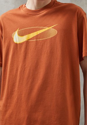 Personne portant un t-shirt couleur rouille avec un logo Nike swoosh jaune à l'intérieur d'un ovale pointillé sur la poitrine.