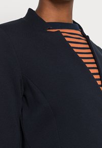 Chaqueta texturizada azul marino con cuello alto, que muestra una camisa a rayas naranja y azul marino en contraste visible debajo.