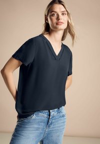 Blusa blu navy a maniche corte con scollatura a V e cuciture dettagliate lungo il colletto, abbinata a jeans azzurri. Tessuto morbido.