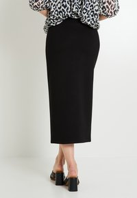 Jupe midi noire ajustée avec une texture lisse, dotée d'une taille haute et d'une fente à l'arrière. Associée à des sandales noires à talons carrés.