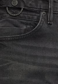 Close-up van een zak van zwarte denim jeans met een metalen knoop en klinknagel met het label "s.Oliver" en een ringtag met de tekst "ELEVATED CASUAL."