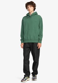 Felpa in cotone verde con cappuccio regolabile e tasca a marsupio frontale, abbinata a jeans neri e sneakers multicolori.