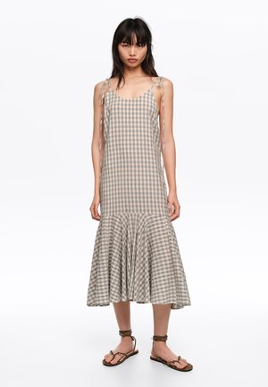 Kobieta w beżowo-szarej, bezrękawnej sukience midi w kratę z wiązanymi ramiączkami i falbaną na dole, w połączeniu z brązowymi sandałami na kostkę z paskiem.