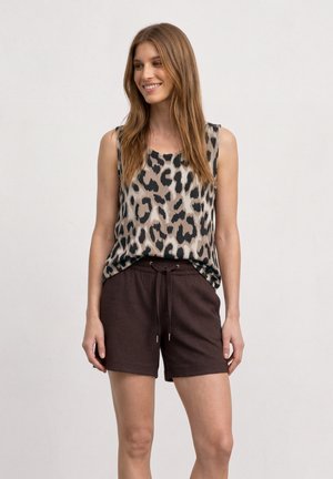 Kvinde med langt brunt hår iført ærmeløs leopardmønstret top og mørkebrune shorts med snøre, smilende og kigger mod højre.