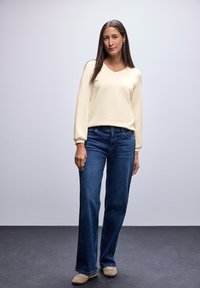 Cremfarbener V-Ausschnitt Langarmtop kombiniert mit dunkelblauen lockeren Jeans und hellbeigen Slippern. Glattes Material und legeres Design.