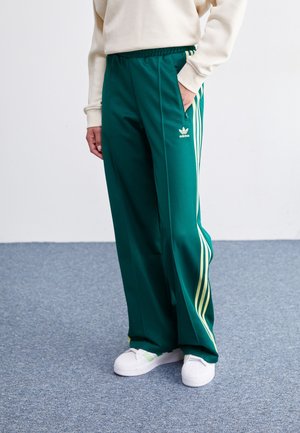Osoba nosząca zielone spodnie dresowe Adidas z białymi paskami i białe buty sportowe, stojąca na niebieskim wykładzinowym podłożu przy białej ścianie.