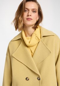 Cappotto giallo doppiopetto in tessuto morbido con ampi revers, abbinato a un dolcevita giallo a coste. Texture liscia e dettagli con bottoni.