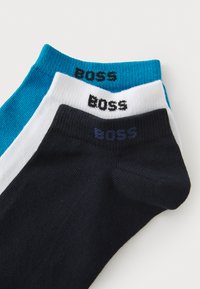 Trois paires de chaussettes basses en bleu marine, blanc et turquoise avec une texture côtelée et le logo "BOSS" imprimé en noir sur chaque chaussette.