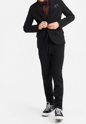 Sort jakkesæt med en tekstureret blazer, maroon skjorte, sort butterfly, tætsiddende sorte bukser og sorte sneakers med hvide såler.