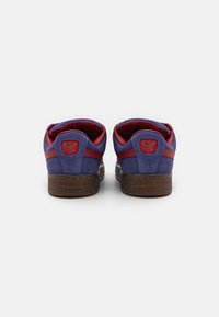 PUMA-sneakers in paarse suede met rode accenten, voorzien van een gestructureerde bruine zool. Gouden logo detail op de hiel. Klassiek lage ontwerp.